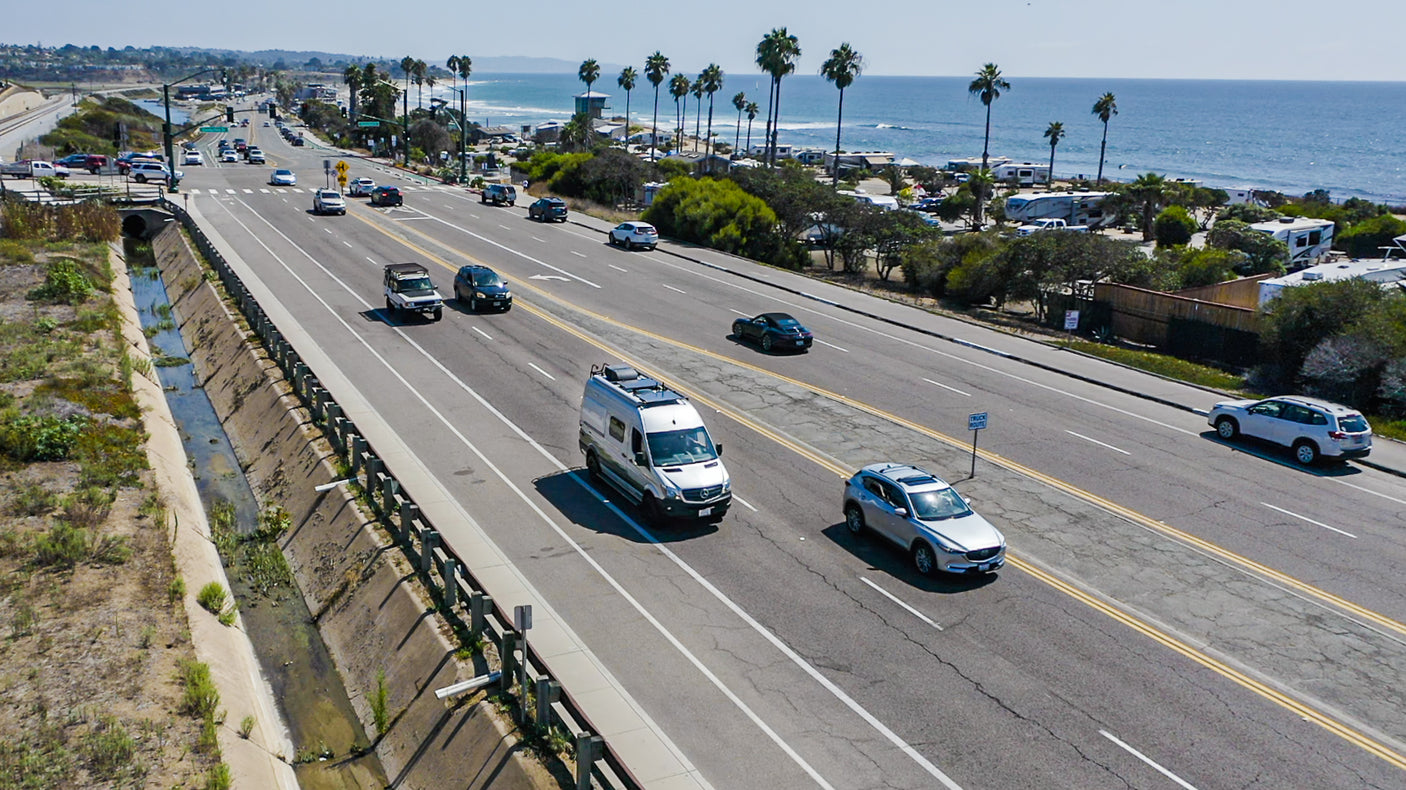 Aerial view: An RV rental drives along a sunny coastal highway by an oceanfront RV park & the Pacific.
