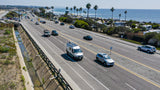 Aerial view: An RV rental drives along a sunny coastal highway by an oceanfront RV park & the Pacific.