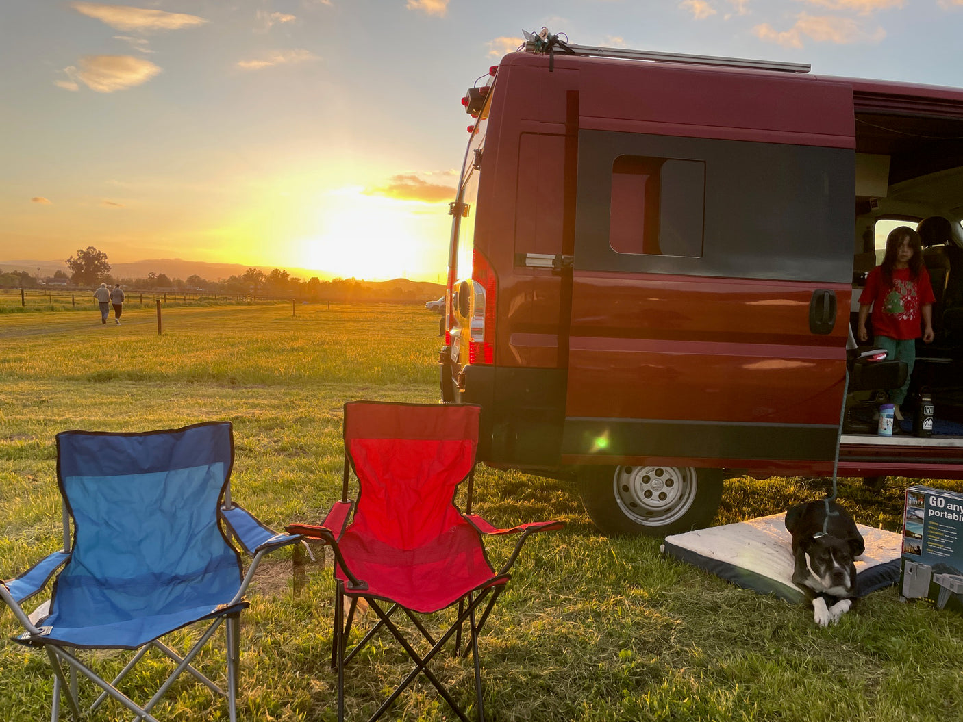 Perfect sunset with an RV rental! Red van, two camp chairs, a dog, and a child on a grassy field.