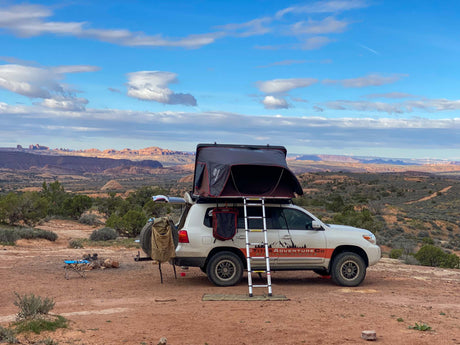 White SUV **RV rental** with rooftop tent & ladder, set for camping in a vast desert landscape under blue skies.