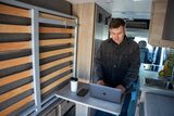 A man works on a laptop in a modern RV rental, featuring a compact kitchen, space-saving bed, and mobile office setup.