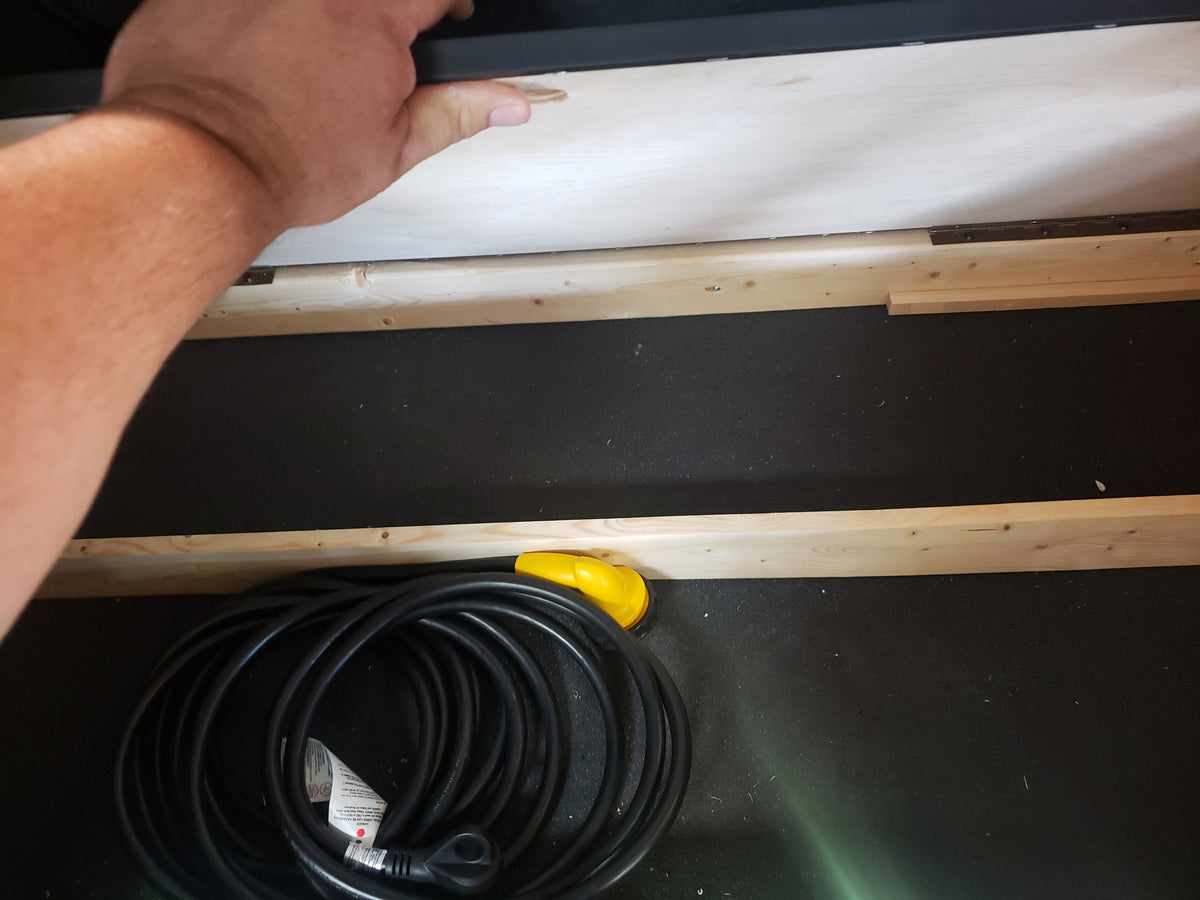 Inside an RV rental storage compartment: a hand reaches over a black power cord, coiled on a dark floor with wood trim. — photo 11