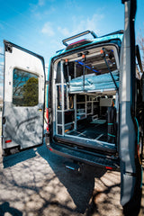 Custom camper van with open rear doors showing modular interior, raised bed, and storage. Ideal for an RV rental.