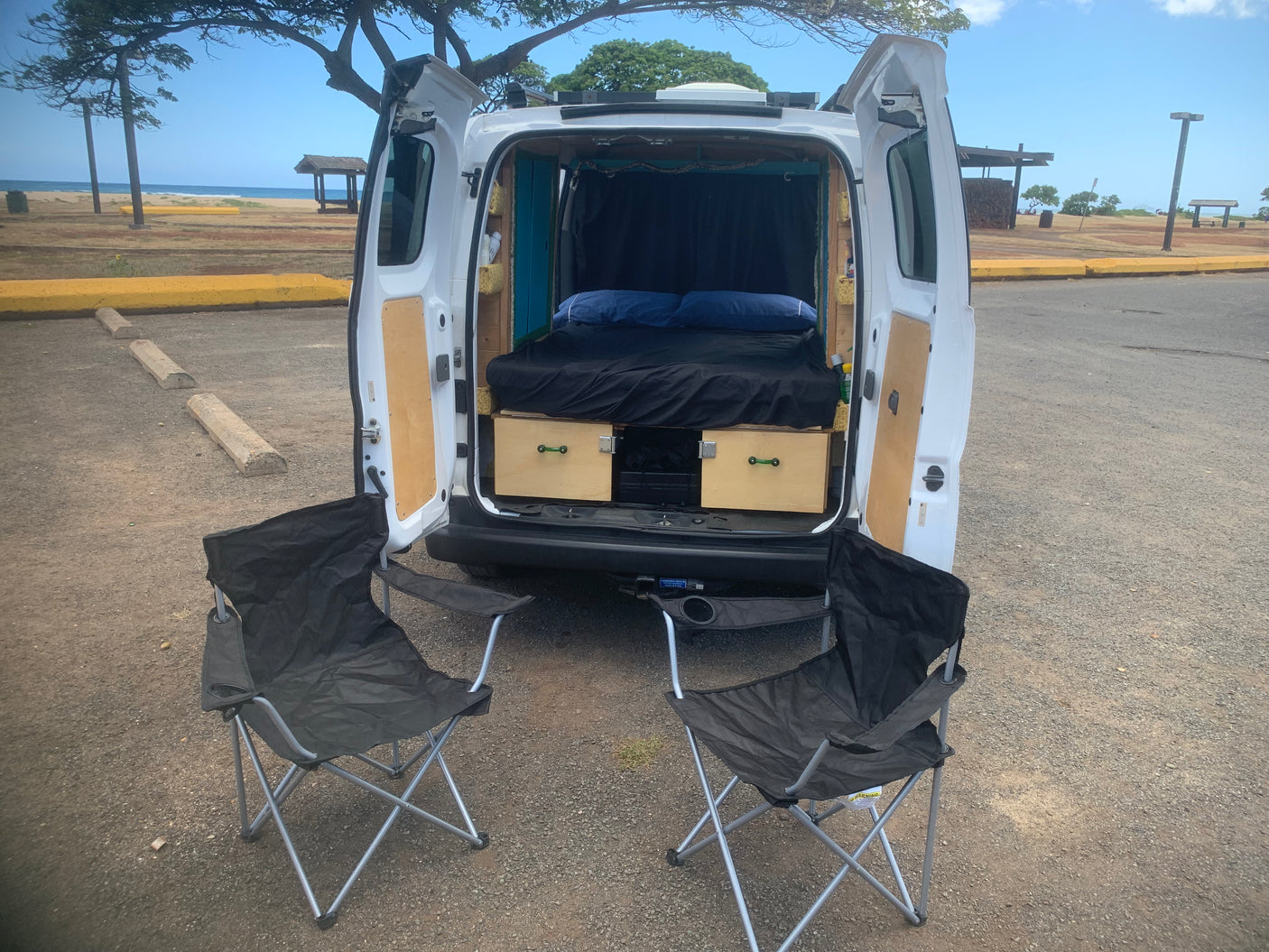 Adventure awaits! This rv rental shows a camper van with open rear revealing a bed, storage, & chairs by a scenic beach.