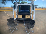 Adventure awaits! This rv rental shows a camper van with open rear revealing a bed, storage, & chairs by a scenic beach.