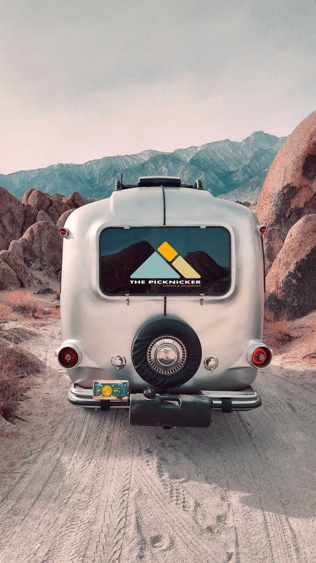 A silver RV rental on a dirt road, framed by mountains & desert rocks. Ready for your next glamping trip!