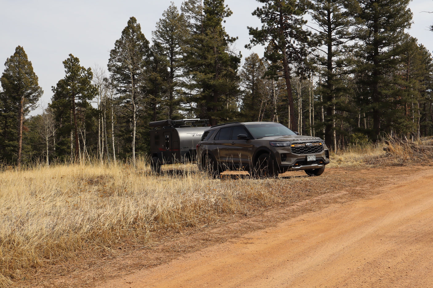 Dark SUV towing a rugged camper trailer on a dirt road through a forest of pines & aspens, perfect for an RV rental adventure.