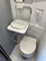 Compact white RV rental wet bath. Features a Thetford toilet, fold-down sink, shower head, and toilet paper.