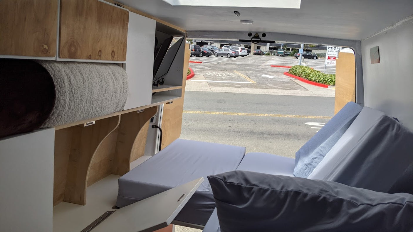 Inside an RV rental camper van: modular wood/white cabinetry, folded grey cushions forming a bed, looking out to a parking lot.