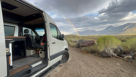 RV rental adventure: Open camper van door shows interior, with a vibrant rainbow over mountains.