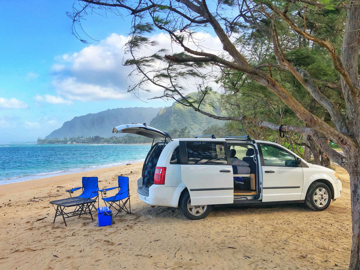 Coastal RV rental adventure: white minivan parked on sandy beach with chairs, overlooking the ocean & mountains.