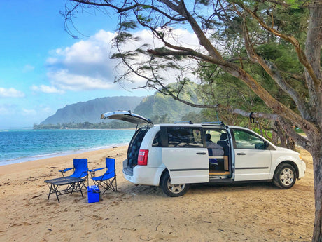 Coastal RV rental adventure: white minivan parked on sandy beach with chairs, overlooking the ocean & mountains.