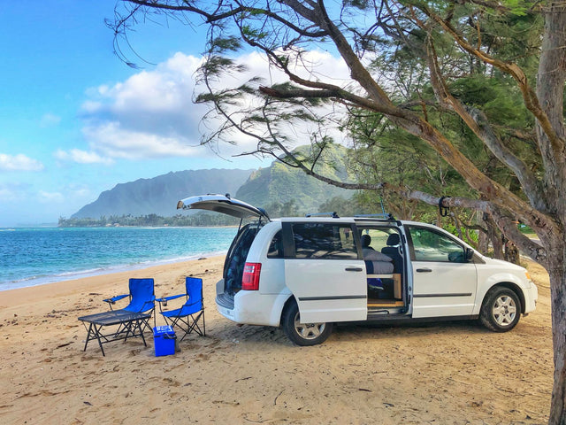 Coastal RV rental adventure: white minivan parked on sandy beach with chairs, overlooking the ocean & mountains.