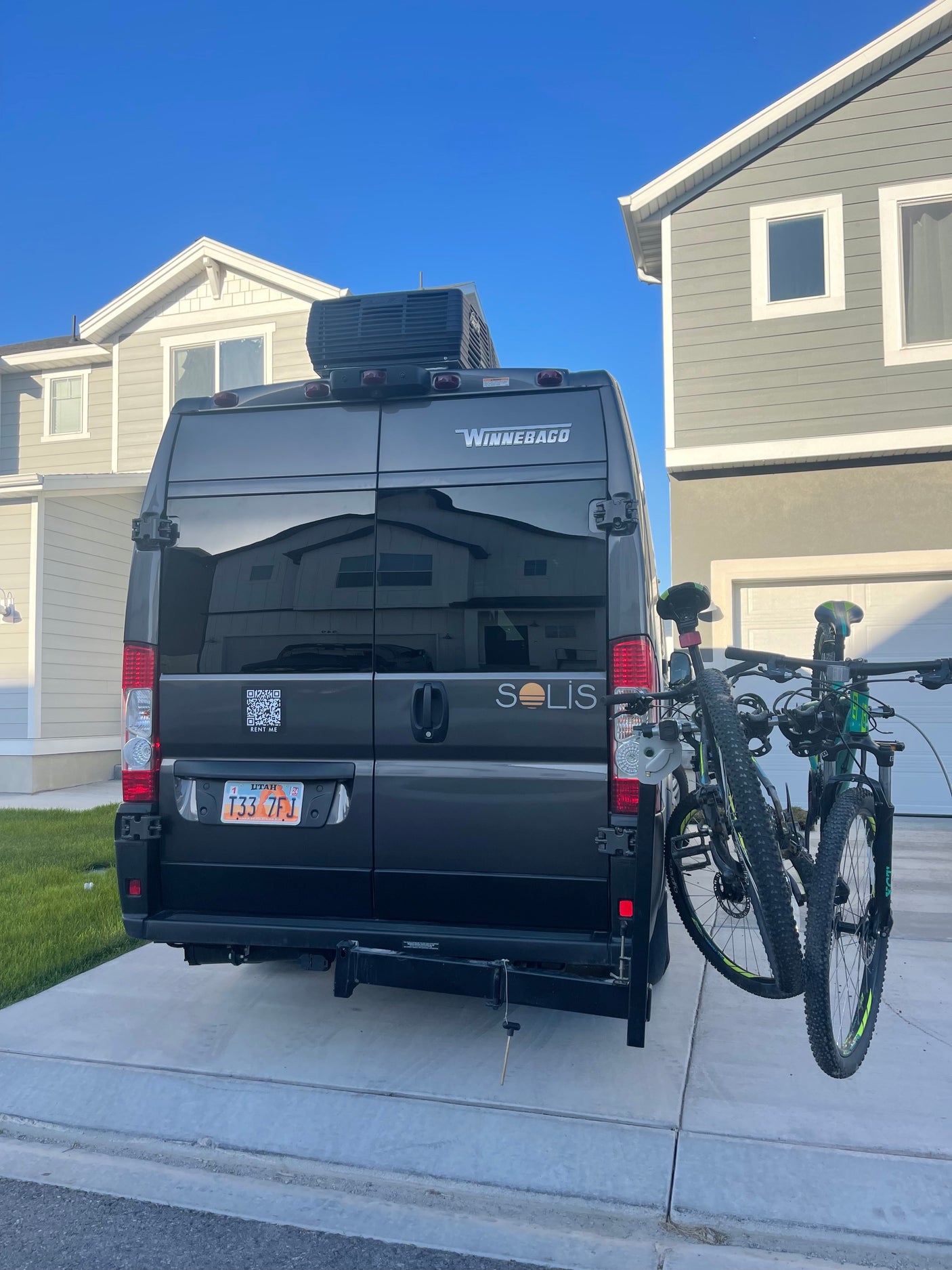 Grey Winnebago Solis **RV rental** van with bikes on a rear rack, parked in a driveway. Utah license plate visible.