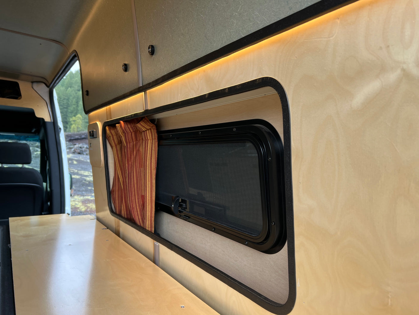 RV rental interior: light wood, ambient lighting, a screened window with a striped curtain, and a bed base for comfort.