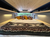 Interior of an **RV rental** featuring a cozy bed with patterned blanket, overhead storage, and nature view.