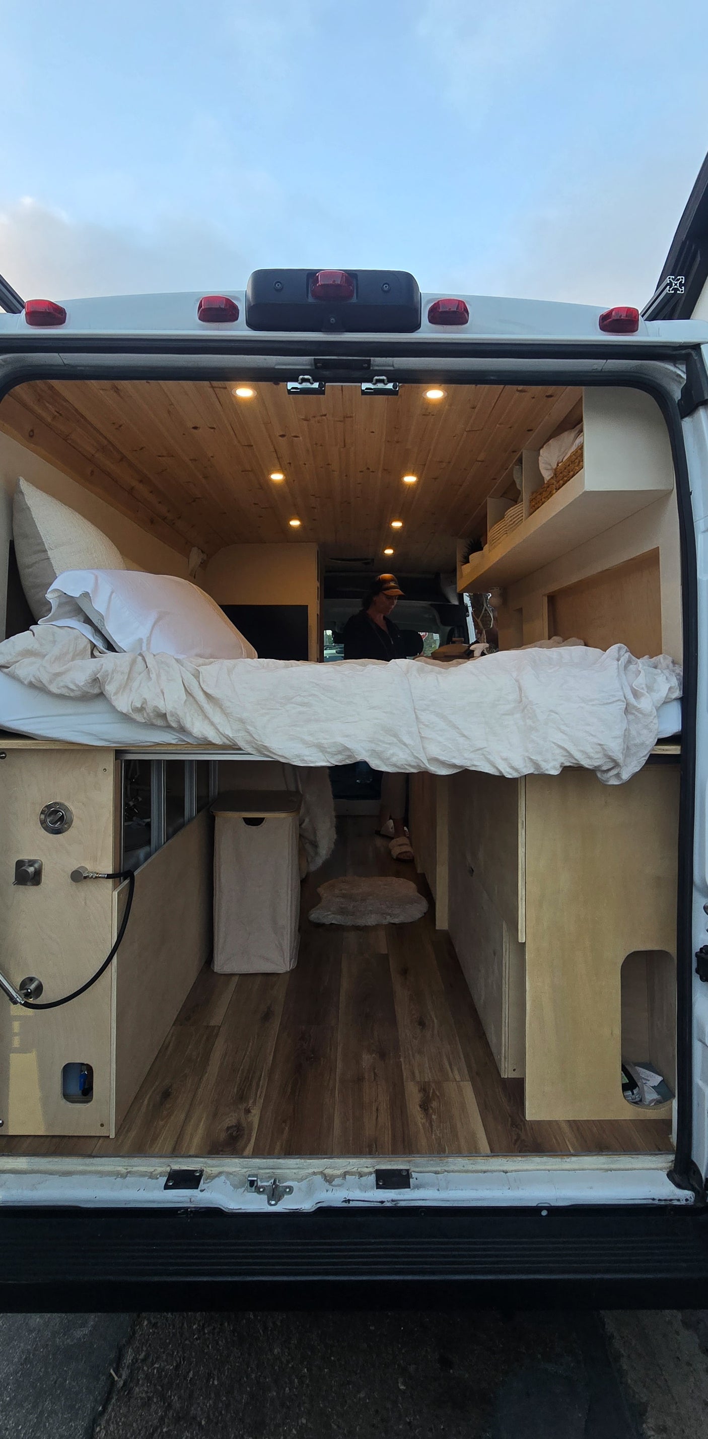 Cozy RV rental interior viewed from the back doors, featuring a raised bed, wooden ceiling, and a person working.