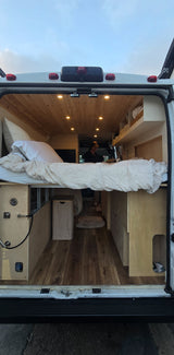 Cozy RV rental interior viewed from the back doors, featuring a raised bed, wooden ceiling, and a person working.