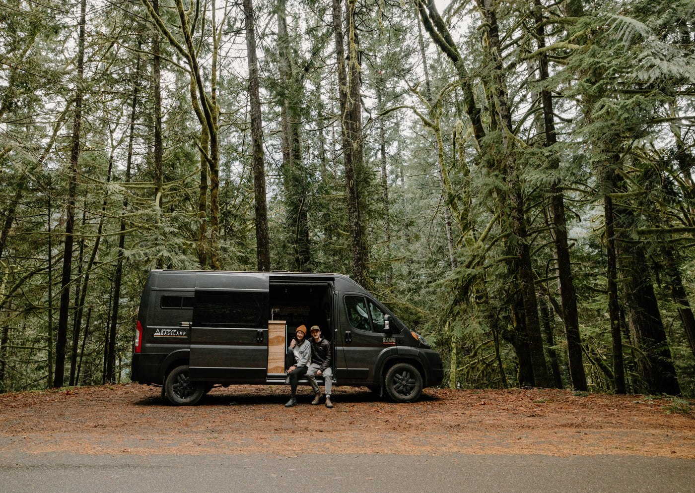 An RV rental parked by a mossy forest allows a couple to enjoy nature from their camper van's open door.