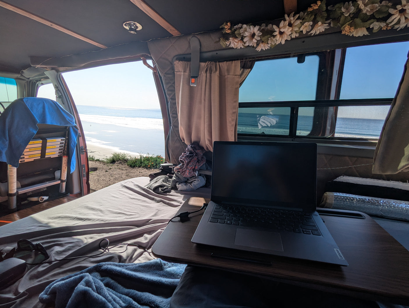 Inside an **RV rental** van, a laptop is set up for remote work on a bed. The open door and window reveal a sunny **beach camping** view.