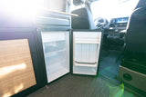 RV rental interior: an open built-in mini-fridge with shelves and door storage. The van's front seats and dashboard are visible.