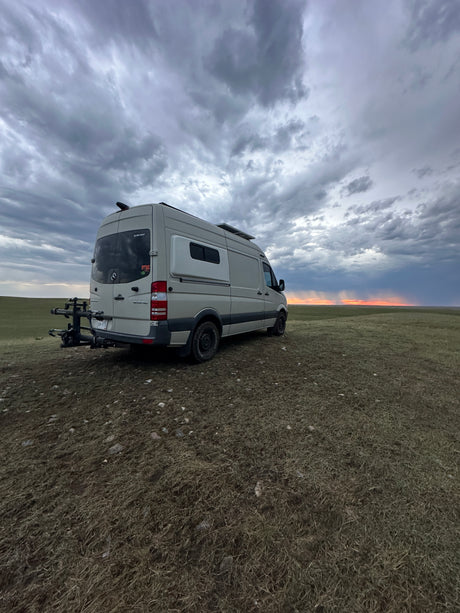 RV rental van parked in a vast field under a dramatic cloudy sky with a sunset horizon. Perfect for adventure!