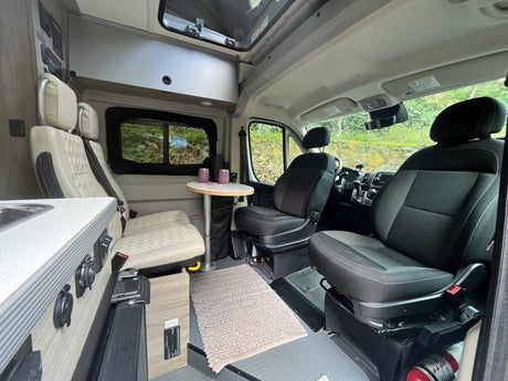 View inside an RV rental showing the living space. Features front swivel seats, a dining area with table & bench seats, and a compact kitchen.