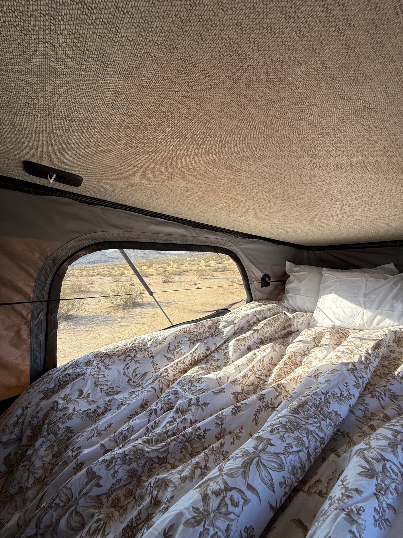 View from inside an RV rental showing a cozy bed with floral duvet, white pillows, and a desert window view.