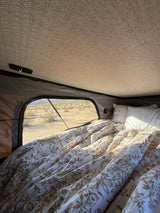 View from inside an RV rental showing a cozy bed with floral duvet, white pillows, and a desert window view.