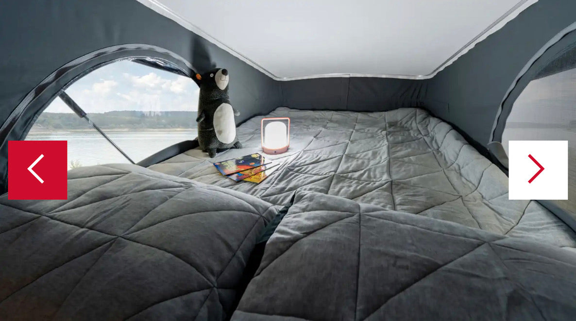 This RV rental offers a cozy camper bed with soft grey blankets, featuring a beautiful lakeside view from the large window.