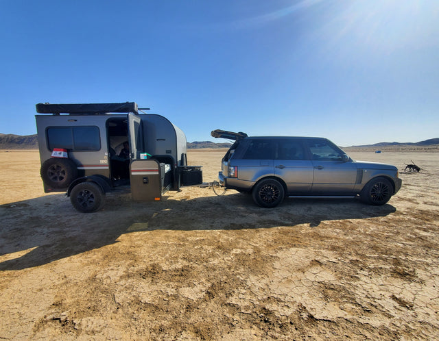 RV rental desert camp: Grey SUV tows a teardrop camper on cracked earth under a clear sky. Ready for adventure!