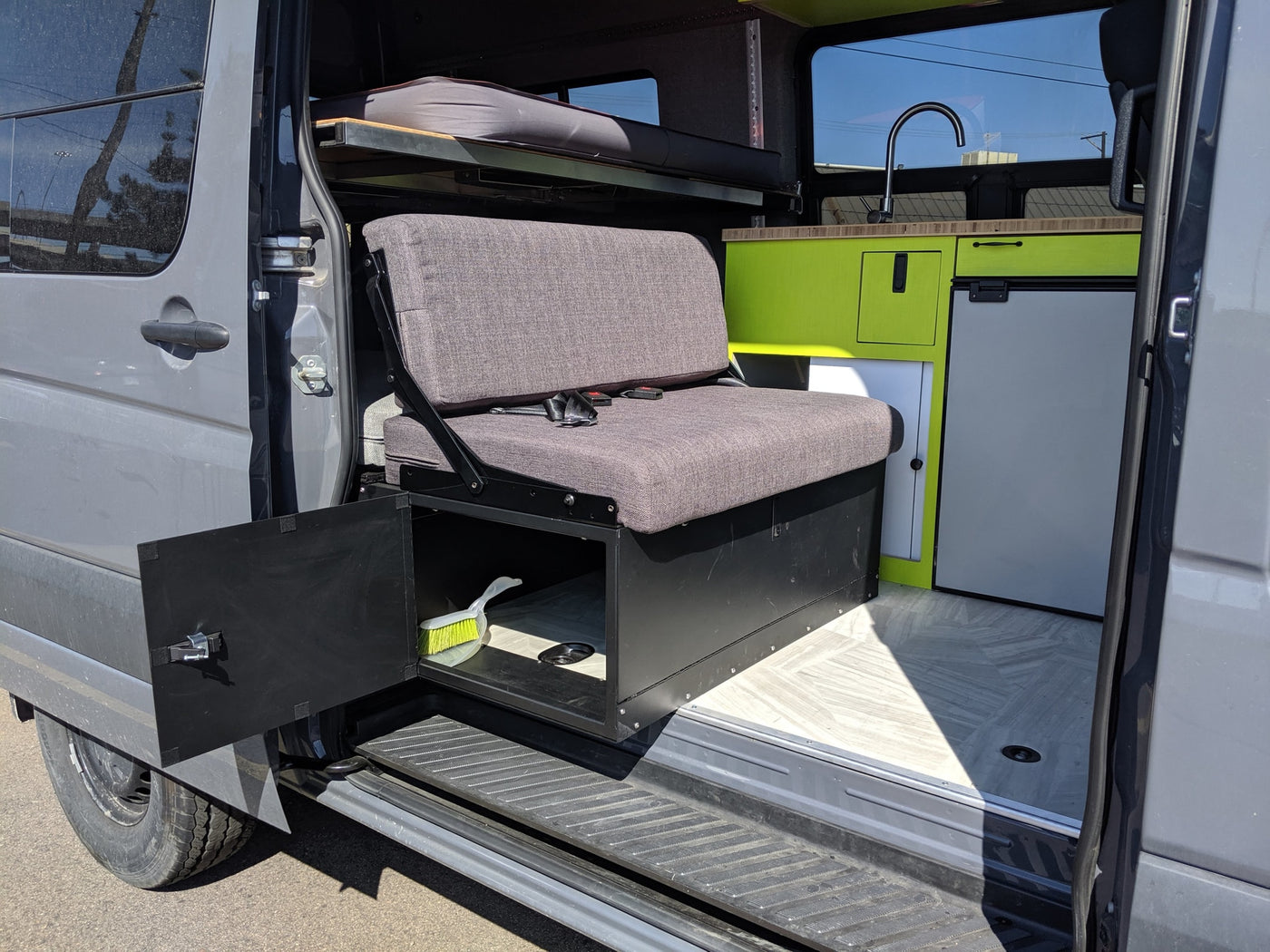 This rv rental van interior shows a grey bench, upper bed, green kitchenette with a sink/fridge, and under-seat storage with a brush. — photo 8