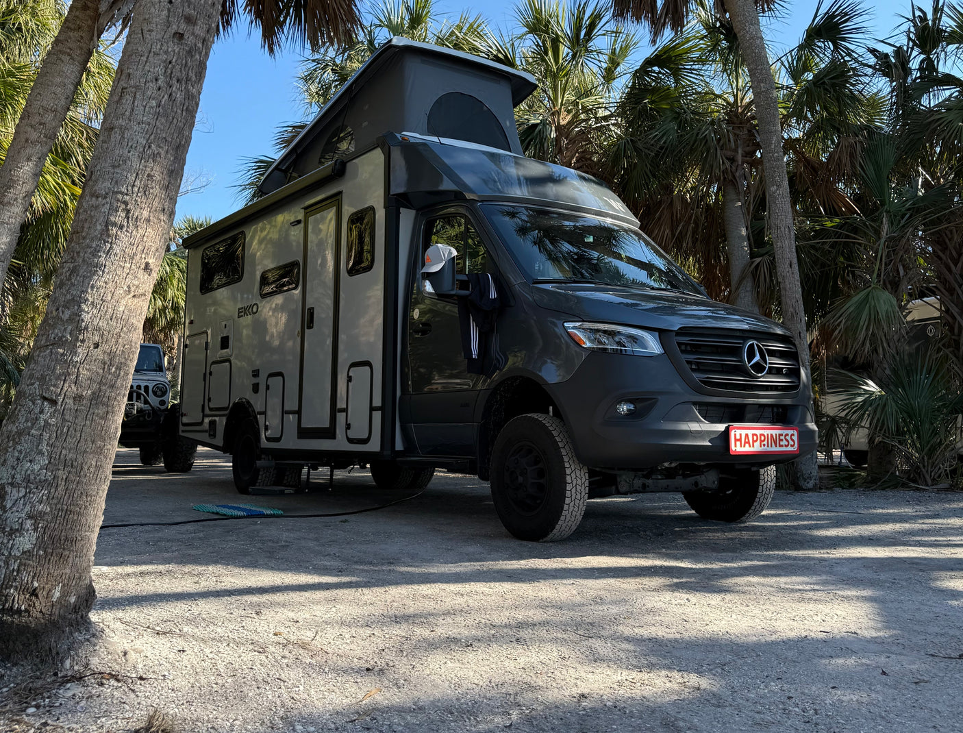 Gray EKKO RV rental (Mercedes camper van) with pop-top, parked on a shell lot under palm trees. Its plate reads HAPPINESS!