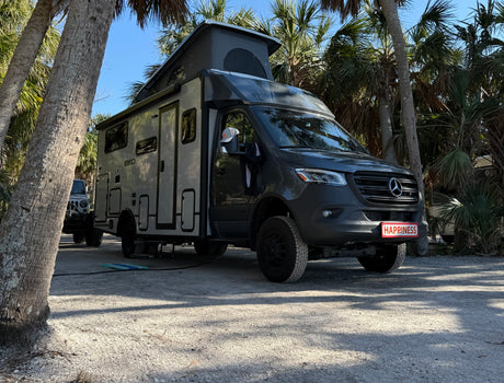Gray EKKO RV rental (Mercedes camper van) with pop-top, parked on a shell lot under palm trees. Its plate reads HAPPINESS!