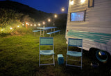 Cozy RV rental setup at night, featuring a vintage camper, string lights, blue picnic table, and chairs on green grass.