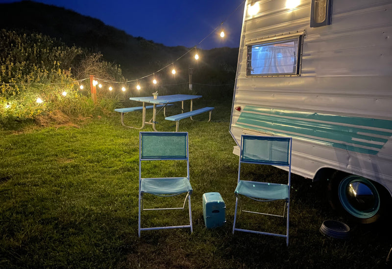 Cozy RV rental setup at night, featuring a vintage camper, string lights, blue picnic table, and chairs on green grass. — photo 2