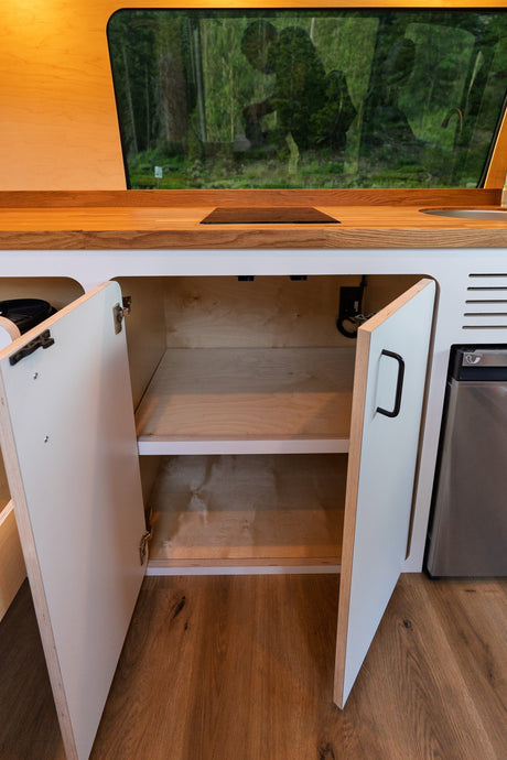 Modern RV rental kitchen inside a camper van, showing wooden counters, an open storage cabinet, and a fridge. Scenic forest view.