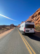 White RV rental van parked on a desert road, amidst stunning red rock formations under a clear blue sky.