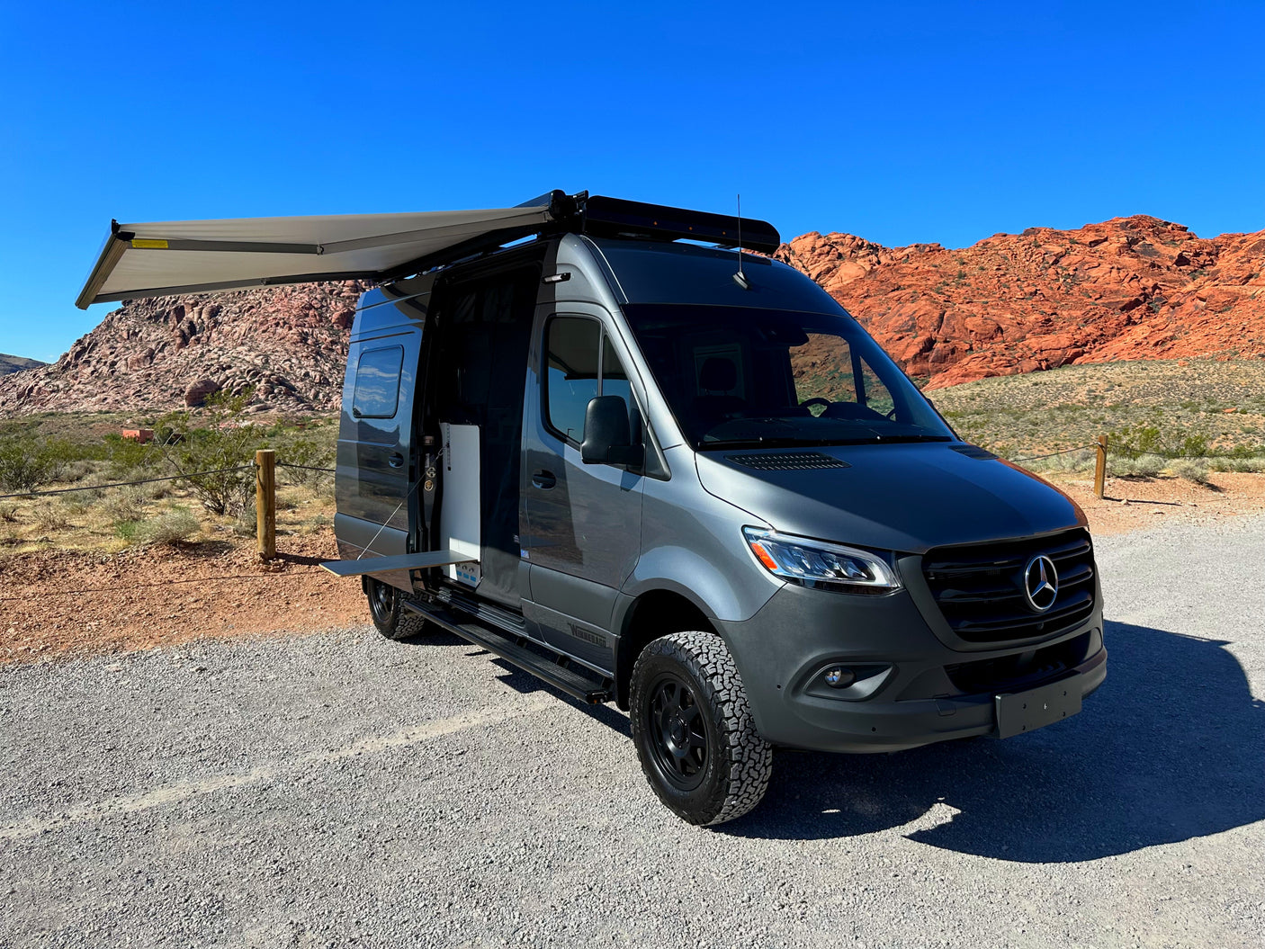 Gray Mercedes Sprinter rv rental van with an extended awning and open side door, parked on gravel with red rock mountains.