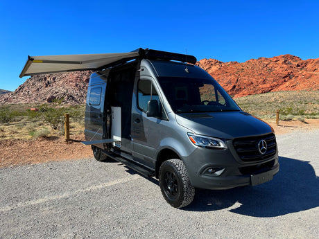 Gray Mercedes Sprinter rv rental van with an extended awning and open side door, parked on gravel with red rock mountains.