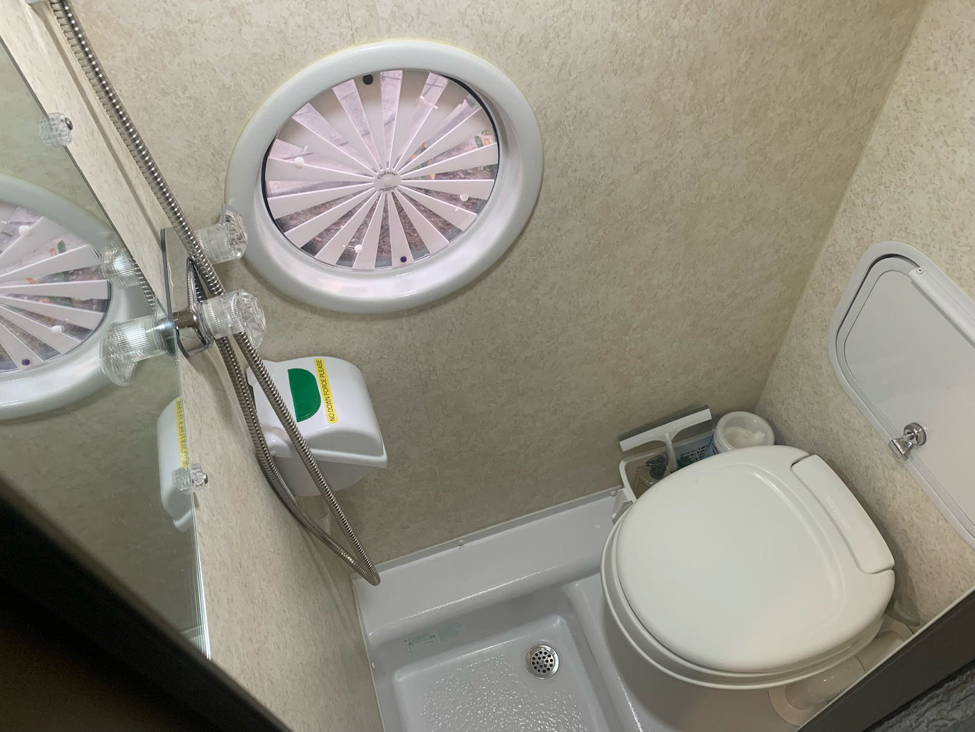 Inside an RV rental wet bath, showing the compact toilet, handheld shower, and an oval overhead vent with slats.