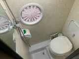 Inside an RV rental wet bath, showing the compact toilet, handheld shower, and an oval overhead vent with slats.