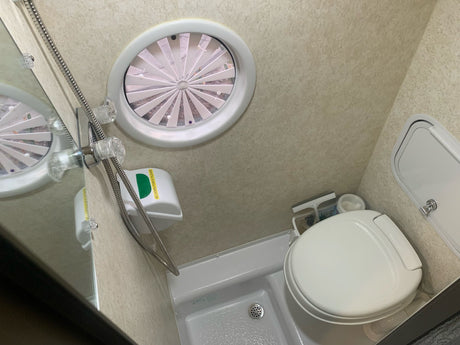 Inside an RV rental wet bath, showing the compact toilet, handheld shower, and an oval overhead vent with slats.
