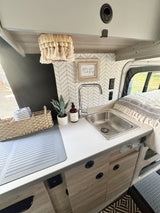 Modern RV rental interior with a cozy kitchenette, including a sink, plant, and decor. Adjacent bed area for comfortable travel.