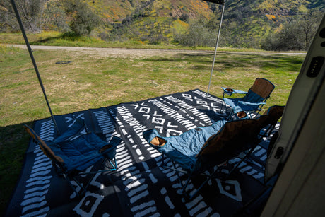 Comfortable RV rental outdoor setup with chairs and patterned mat, facing a green, flower-speckled hill.