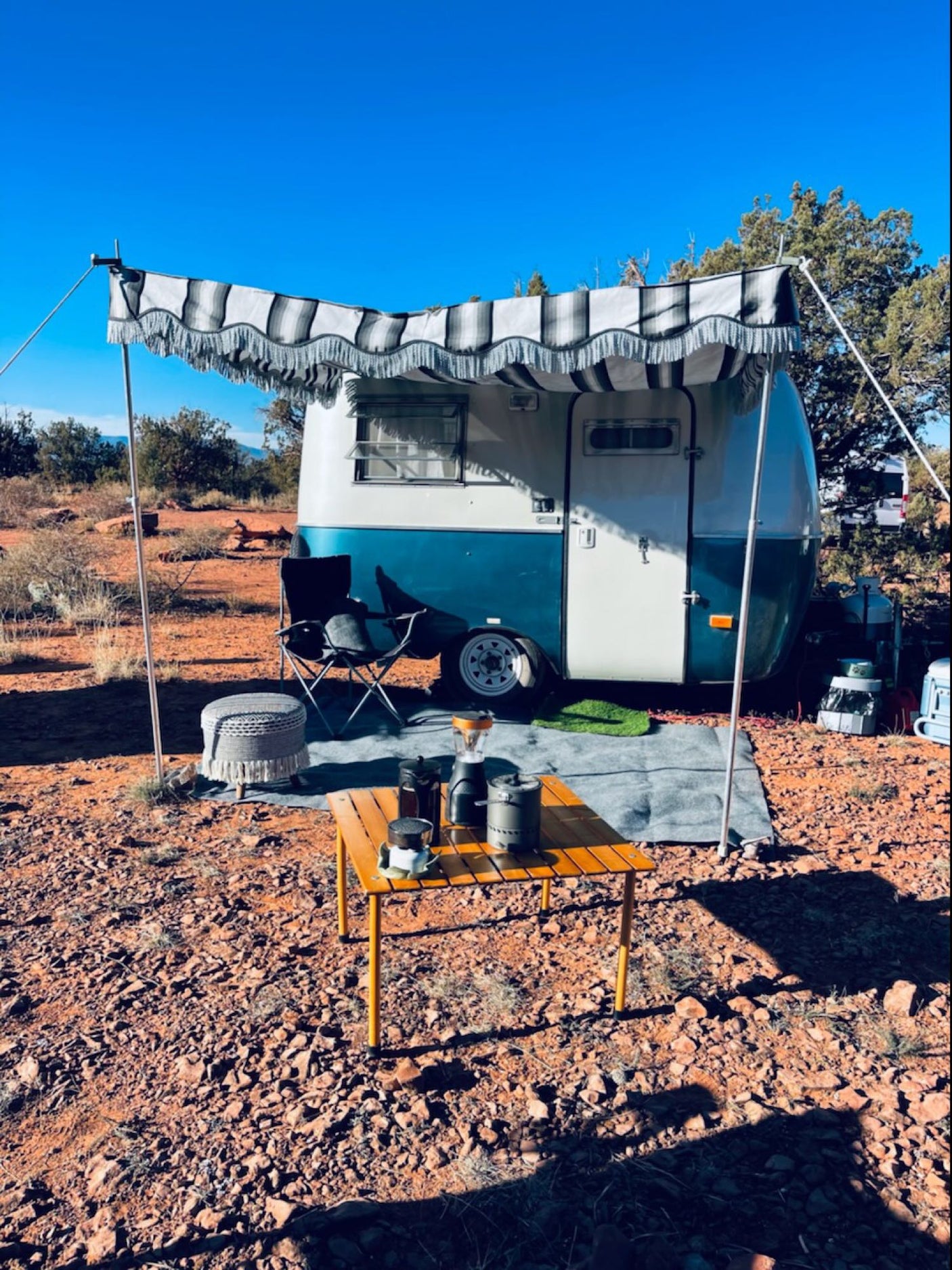 This RV rental shows a retro camper with an awning & coffee setup, amidst a rocky outdoor camp.