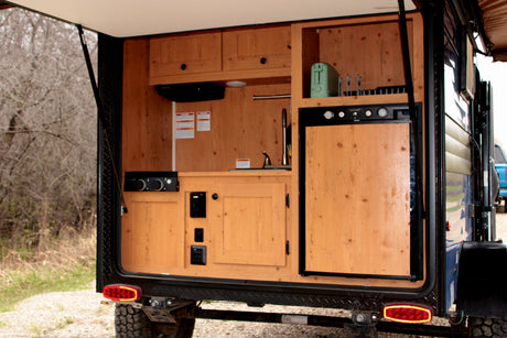 RV rental: Rear galley of a camper trailer featuring a wooden outdoor kitchen with a fridge, sink, and toaster, ready for adventure.