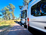 Rear view of a white Coachmen camper van RV rental with a bike rack, parked among pine trees for camping.