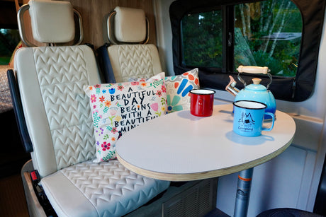 This RV rental interior shows plush seating, a table with a kettle and mugs, ideal for comfortable travel and camping.