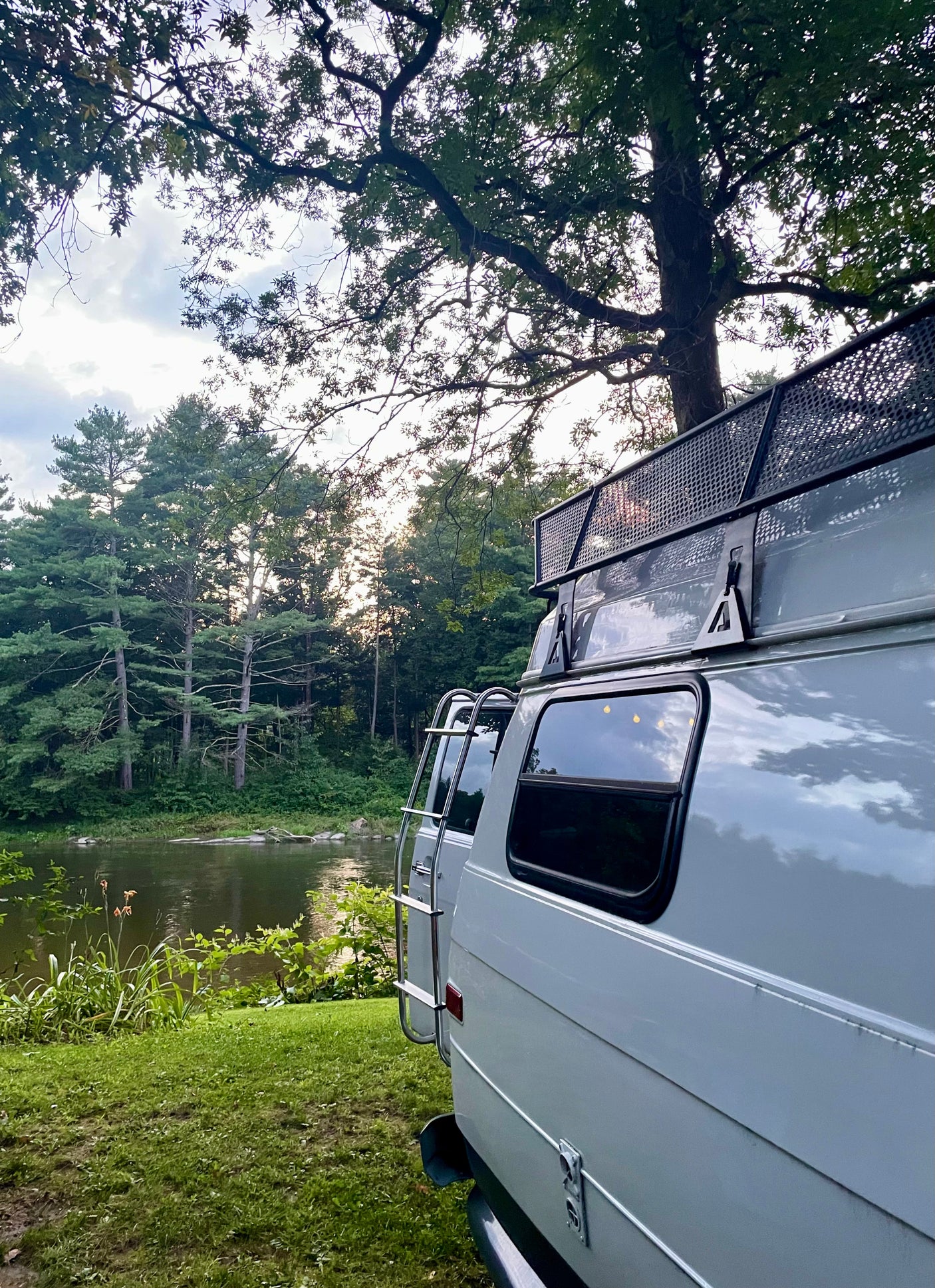 Scenic **RV rental** view: a white van, equipped with roof rack and ladder, parked by a tranquil river amidst lush trees.